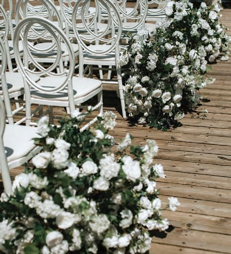 White chairs lined along a wooden aisle with cascading white rose arrangements at the edges, as in a wedding setting.