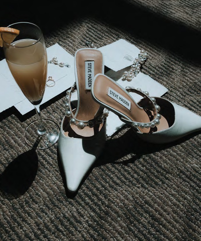 White Steve Madden high-heeled sandals with pearl-embellished straps on a textured carpet, beside a champagne glass and napkins.