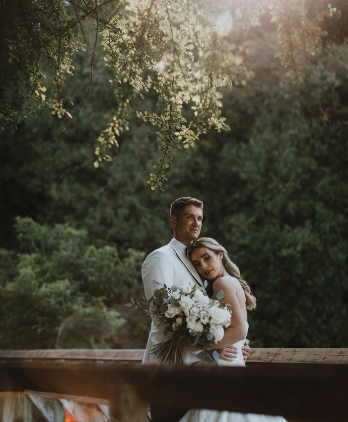 Bride and groom embrace on a wooden railing outdoors, holding a bouquet of white flowers under sunlit foliage.