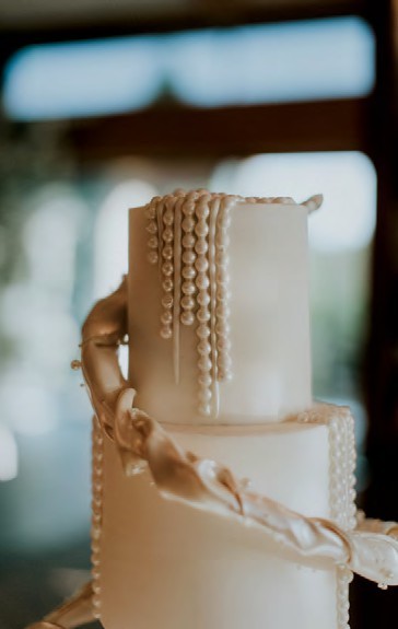 Two-tier white wedding cake with pearl bead garlands cascading down the sides and a braided ribbon detail between tiers (soft-focus background).
