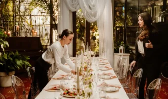 Two women set a long formal dining table with candles, flowers, and glassware in a bright indoor venue.
