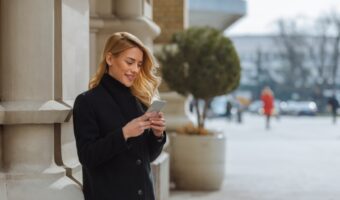 Woman in a black coat stands outside a building, looking at her smartphone.
