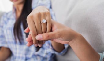 Close-up of a hand with a diamond engagement ring being held by another person’s hand on a soft background.