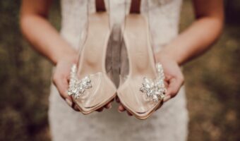 Beige satin high-heeled bridal shoes with crystal floral embellishments held in front of a wedding dress.