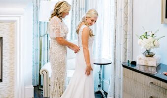 Bride in a white wedding gown stands in a bright room while her mother adjusts the back of her dress.