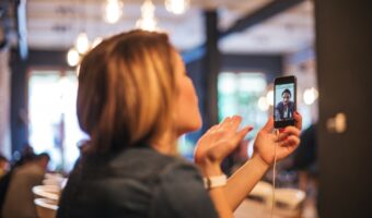 Woman in a café holding up a smartphone for a video call, the caller’s face visible on the screen.