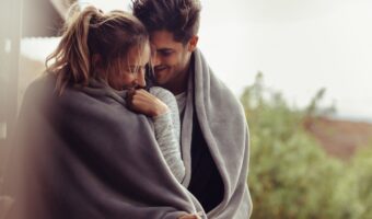 A couple snuggled in blankets outdoors, smiling and leaning close to each other.