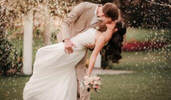 Groom dips and kisses his bride outdoors as confetti falls around them during their wedding.