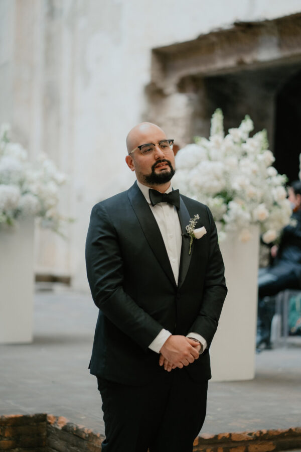 Bald man in a navy tuxedo with a black bow tie and boutonniere, standing with hands clasped at a wedding, with white flowers in background.
