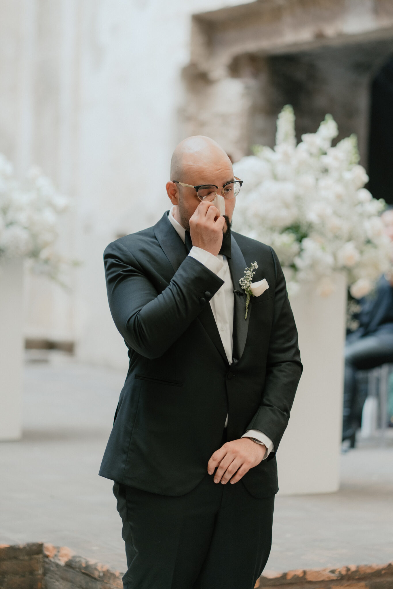 Man in a dark suit wipes his nose with a tissue at an outdoor wedding-like setting, glasses on, floral backdrop behind him.