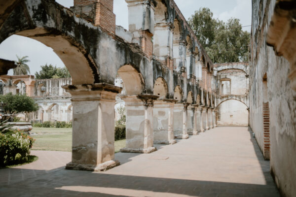 Ruined stone arcade with tall brick arches along a sunlit courtyard; trees visible in the background in the distance.