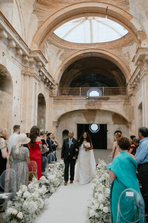 Bride in a white wedding gown walking arm-in-arm with a groom down an aisle filled with white flowers in a historic arched venue.