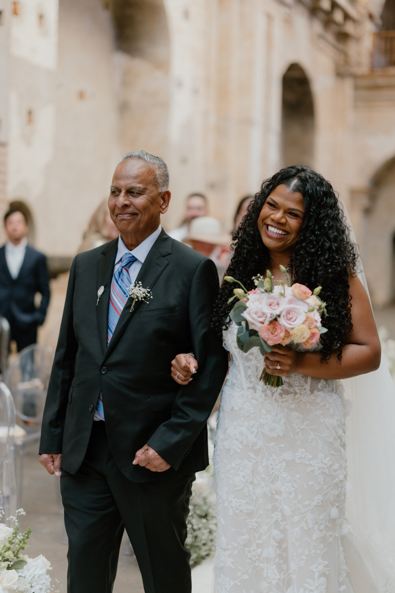 Bride in a white lace wedding gown smiles while walking arm-in-arm with a man in a dark suit, holding a bouquet of pink roses and greenery.