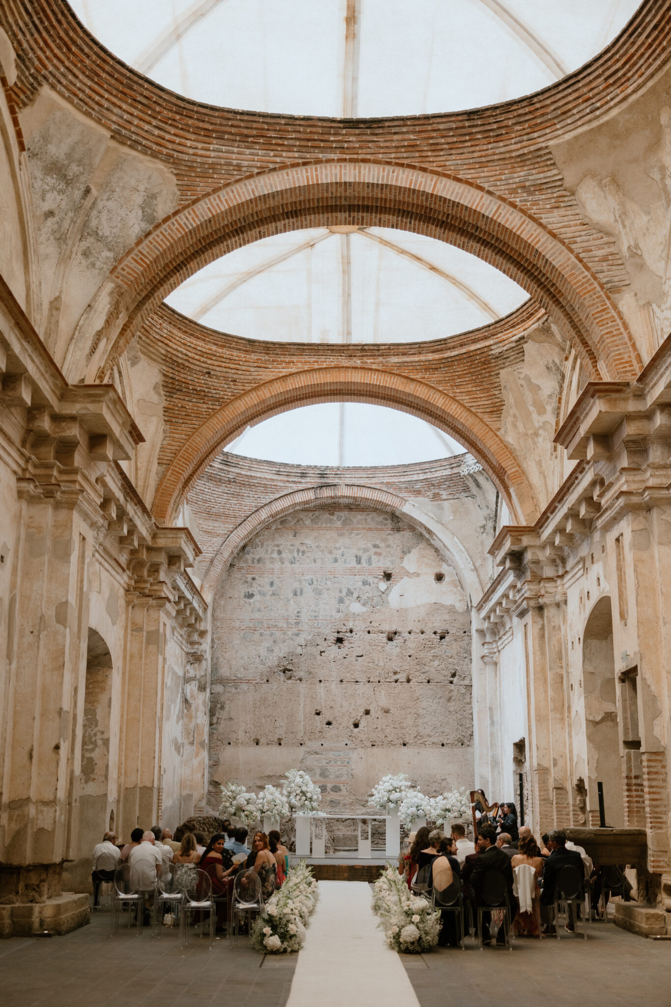 Wedding ceremony set in a historic ruin with an aisle, white flowers, and guests seated on both sides beneath brick arches.