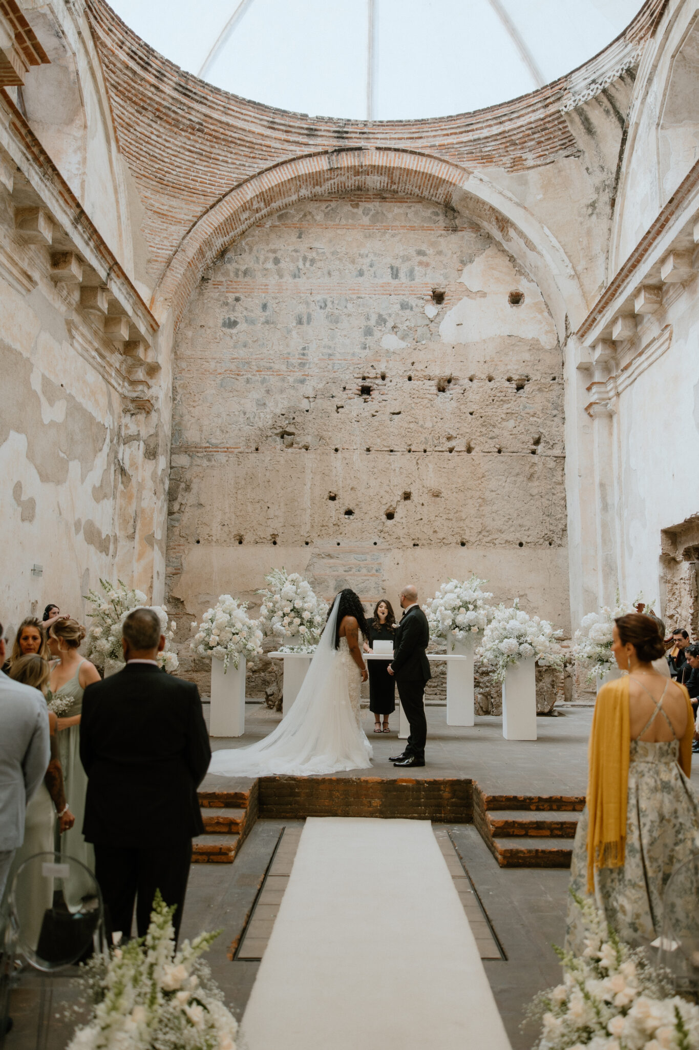 Bride and groom stand at the altar with officiant in a ruined brick church, surrounded by floral arrangements and guests sitting along a white flower-lined aisle.