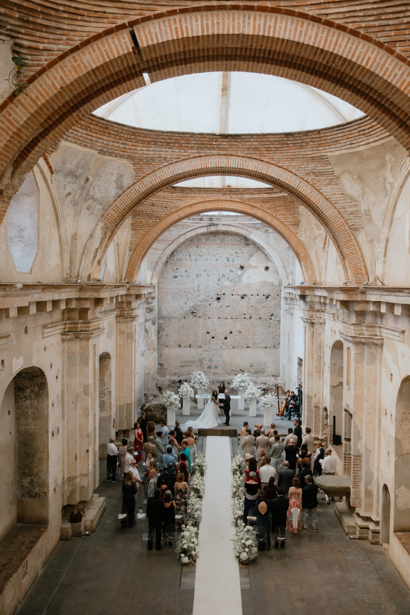 Bride and groom exchange vows at the end of a white aisle lined with white floral arrangements inside a brick arch hall with guests seated on both sides.