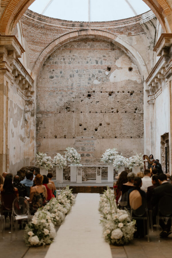 Wedding ceremony in a historic brick hall with a white floral aisle and guests seated along both sides.