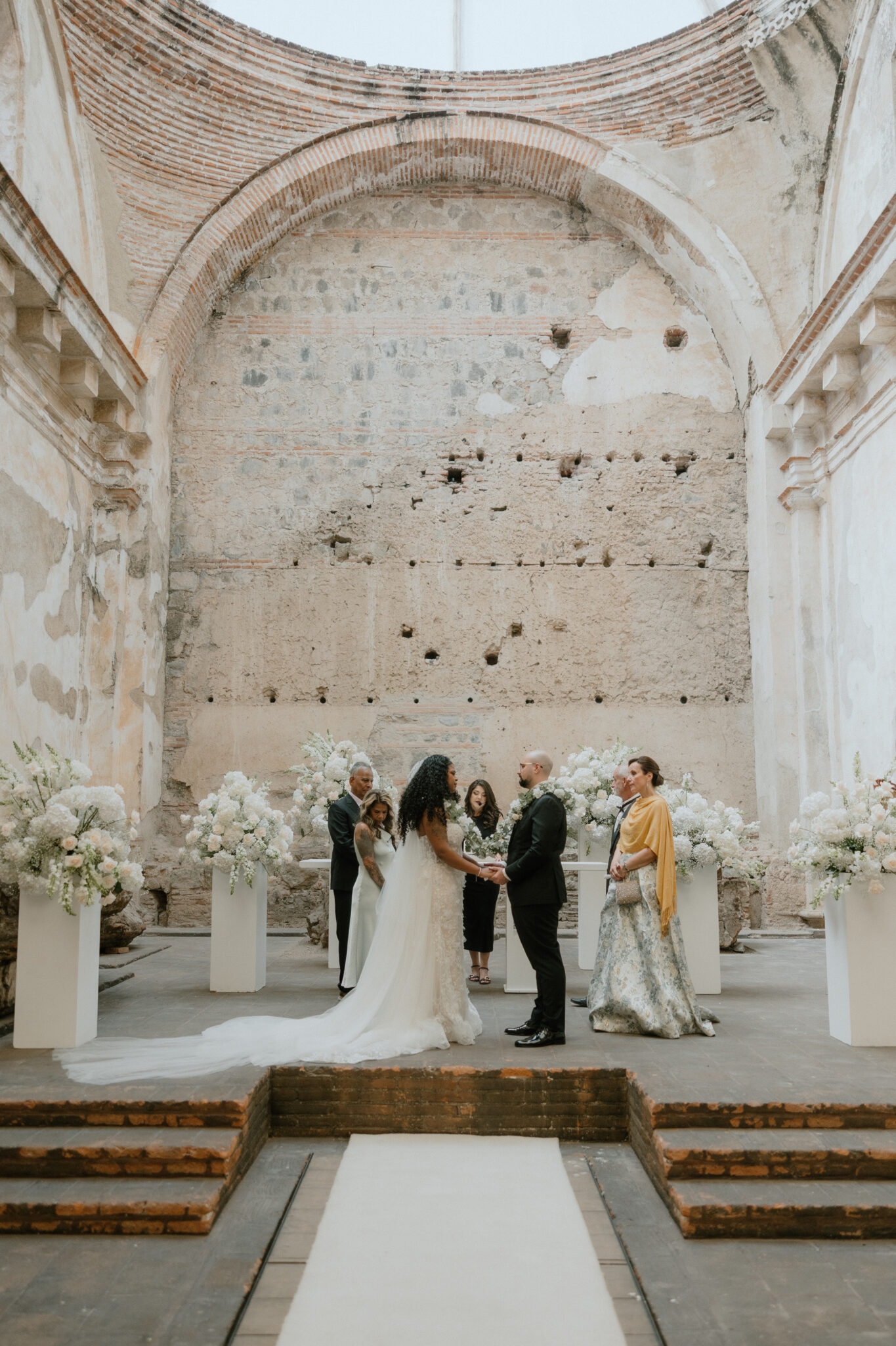 Bride and groom exchange vows in a ruin-like hall with guests and tall white flower arrangements along a white aisle runner.