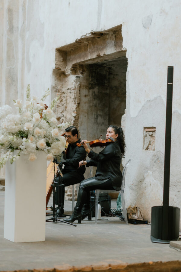 Two violinists in black perform a duet in a ruined whitewashed wall setting with pale floral arrangements nearby.