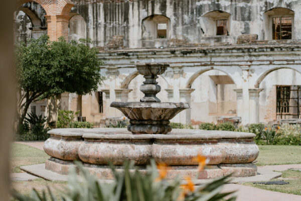 Stone multi-tier fountain in a sunlit historic courtyard with weathered arches and a crumbling facade in the background, greenery nearby.