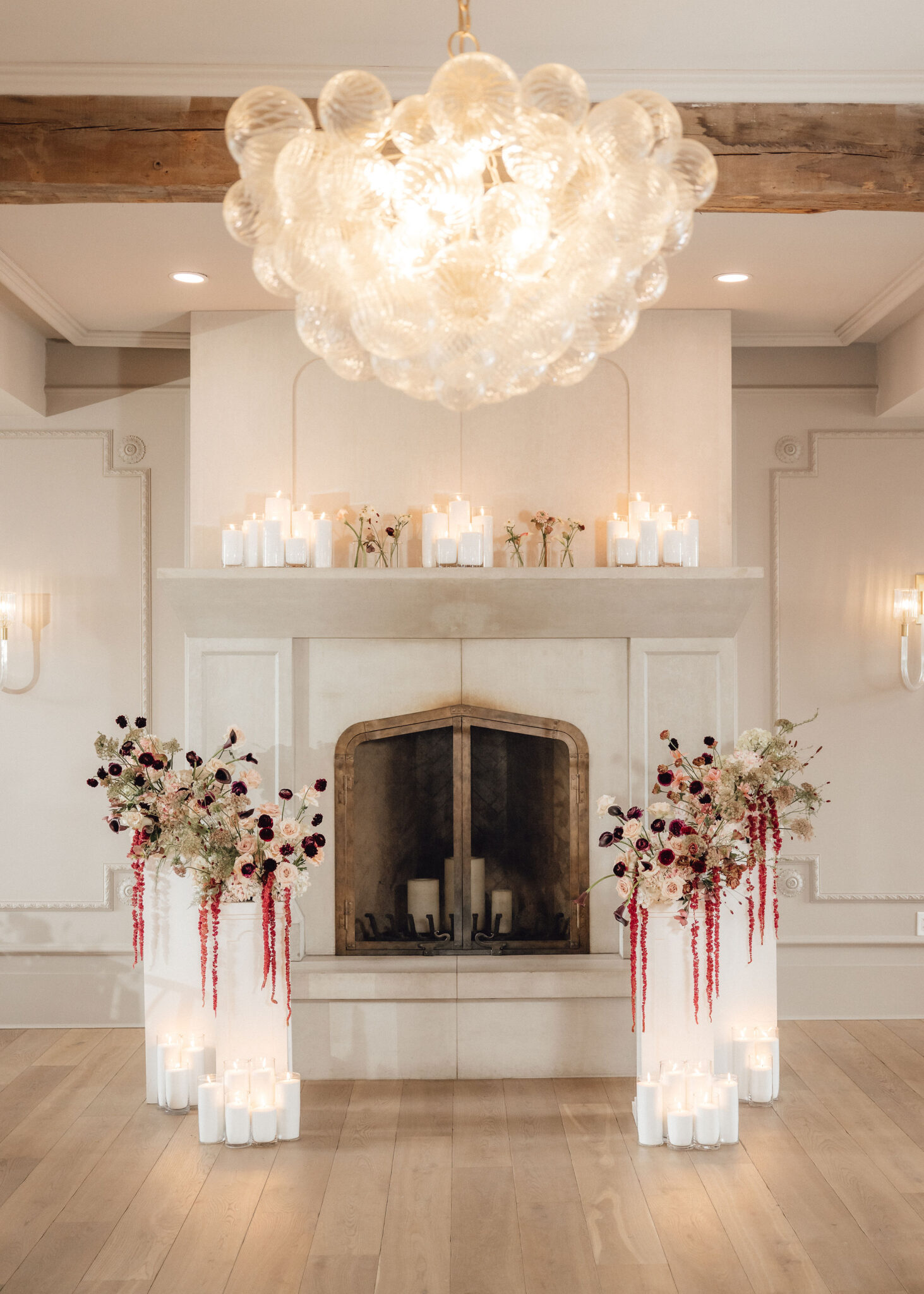 Elegant fireplace setup with tall floral arrangements, white candles, and a crystal chandelier overhead.