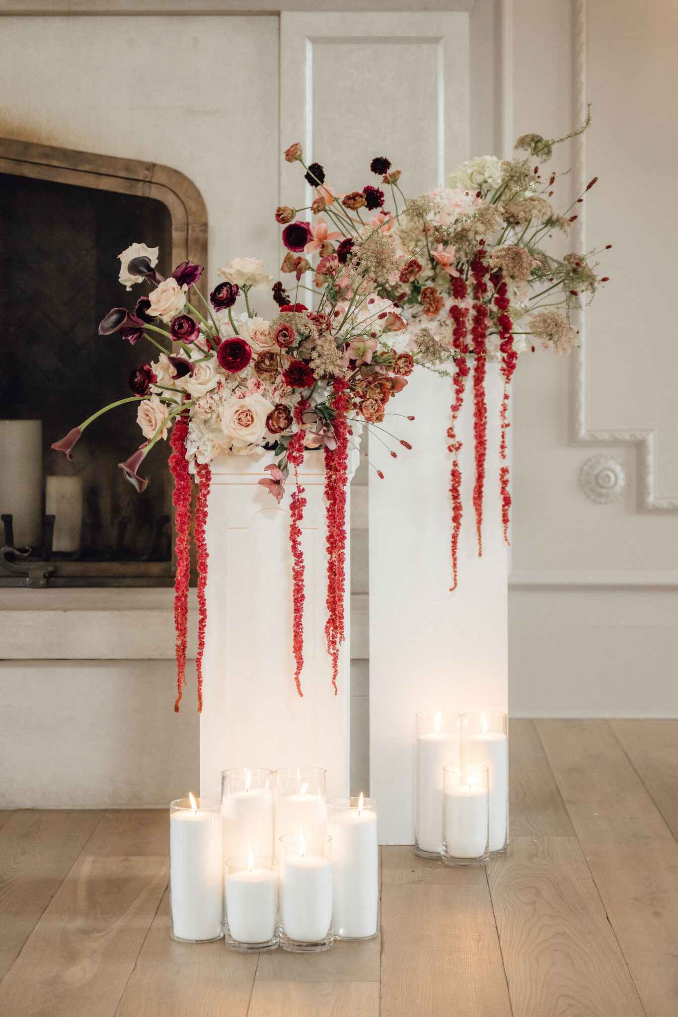 Elegant floral arrangements in pink, white, and burgundy cascading from white pedestals, with trailing red amaranthus and lit candles on the floor nearby in a softly lit room.