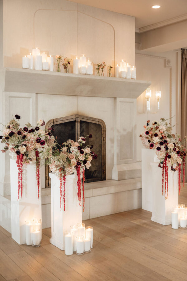 Elegant fireplace mantel decorated with numerous white pillar candles and delicate pink-cream floral arrangements for a chic event setting, with candles in glass holders on the floor nearby.