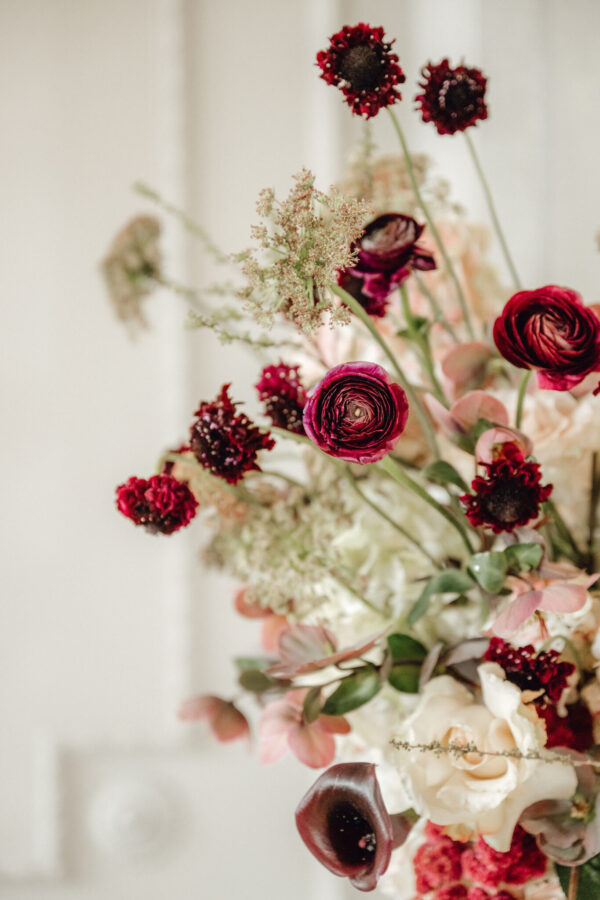 Close-up of a florist bouquet with deep red ranunculus, burgundy blooms, and pale pink and cream flowers with green foliage in soft focus.
