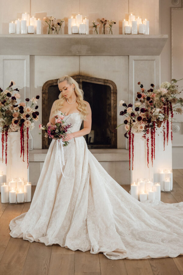 Bride in a white lace gown holding a bouquet, standing before a candlelit fireplace with tall floral arrangements on either side.