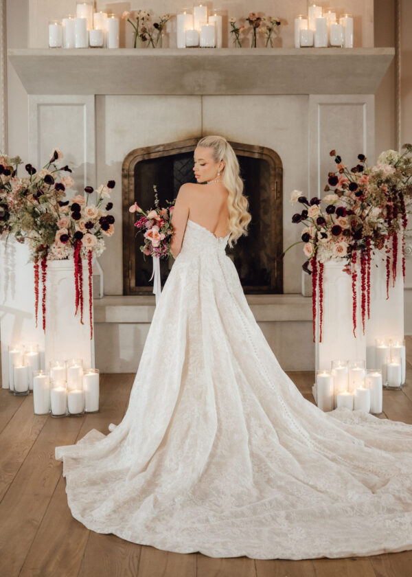 Bride in a white lace wedding gown with a long train standing before a fireplace, holding a pink bouquet among candlelit florals.