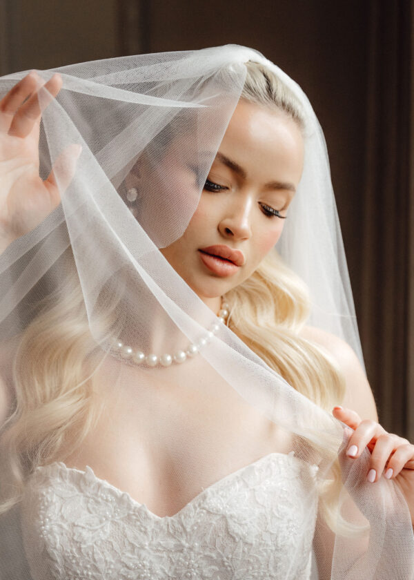 Bride wearing a lace wedding dress and pearl necklace, veil draped over her face.