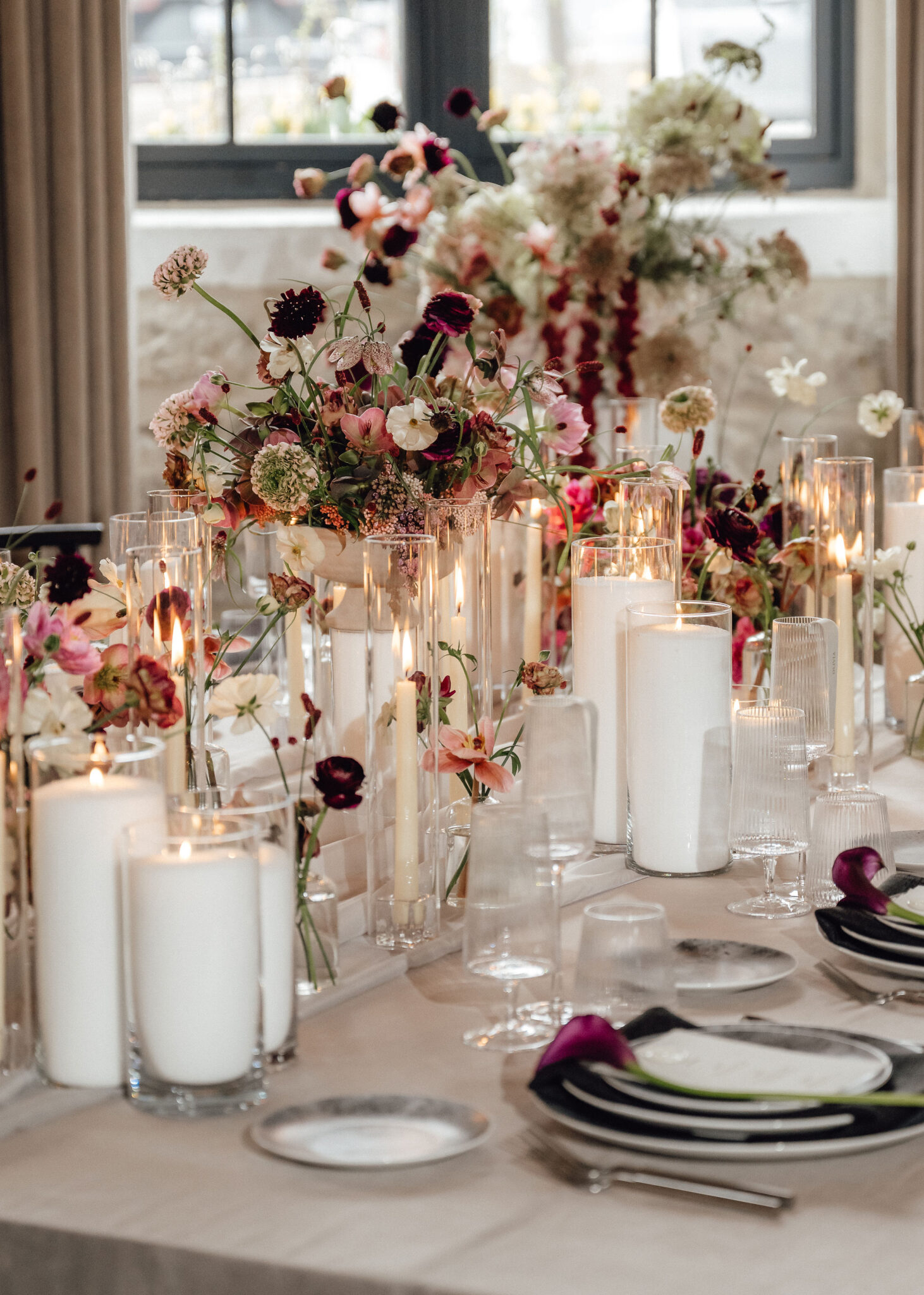 Elegant banquet table with tall white candles, crystal glassware, and lush pink and burgundy flower arrangements as a centerpiece.