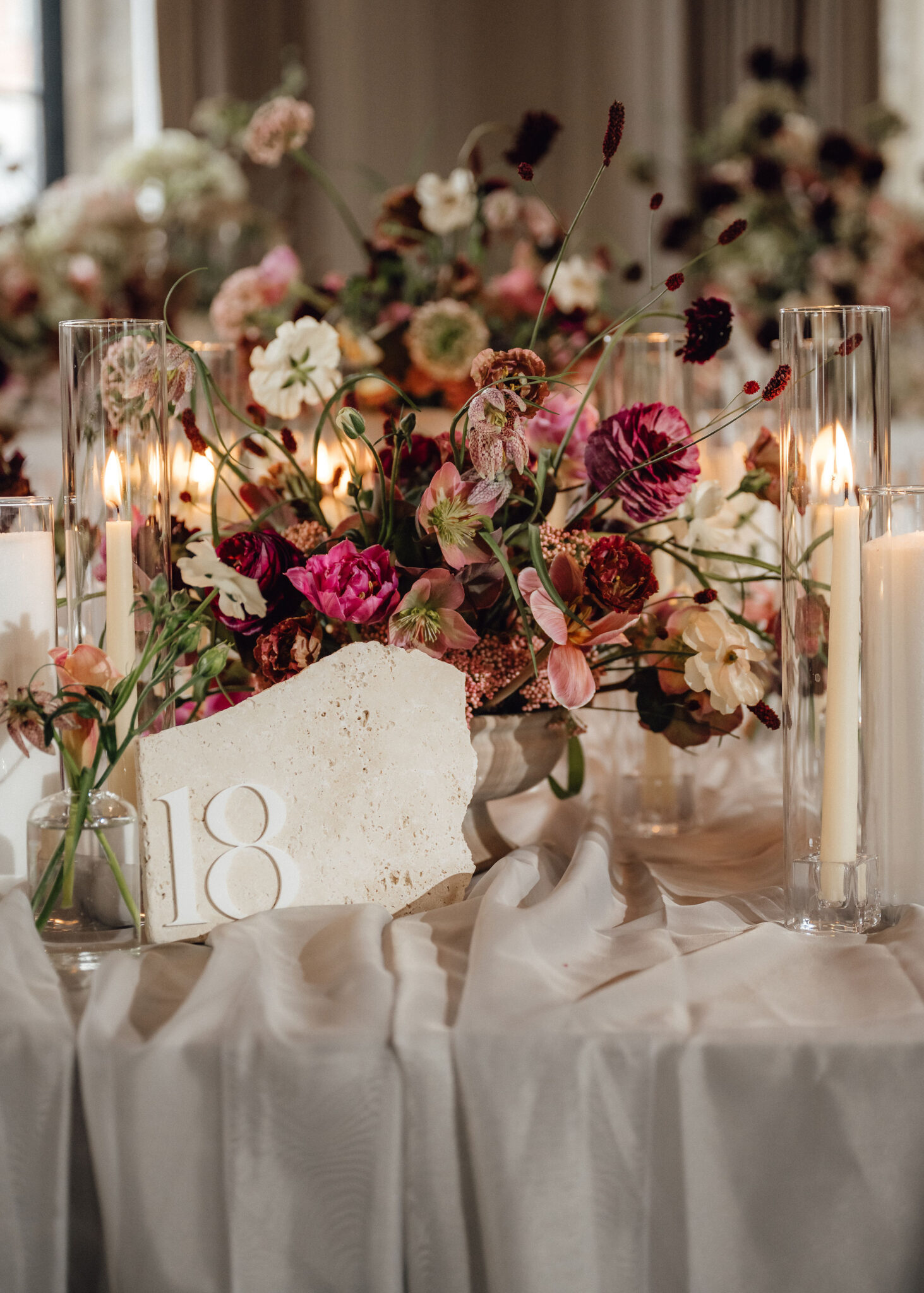 Elegant wedding centerpiece with a lush mix of pink, burgundy, and cream flowers surrounded by tall glass candles on a white tablecloth.