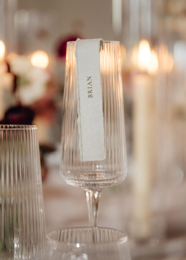 Crystal champagne flute with a white torn-paper tag reading BRIAN, warm candlelight bokeh in the background.