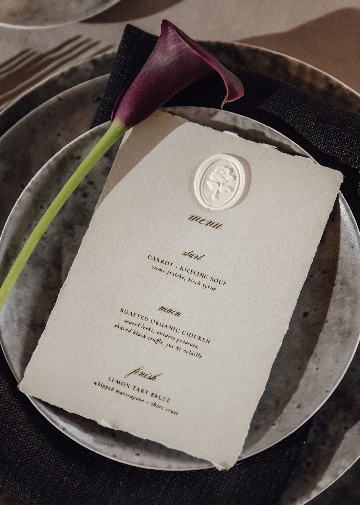 Close-up of an elegant place setting: a cream menu card with a white wax seal rests on a plate, alongside a purple calla lily.
