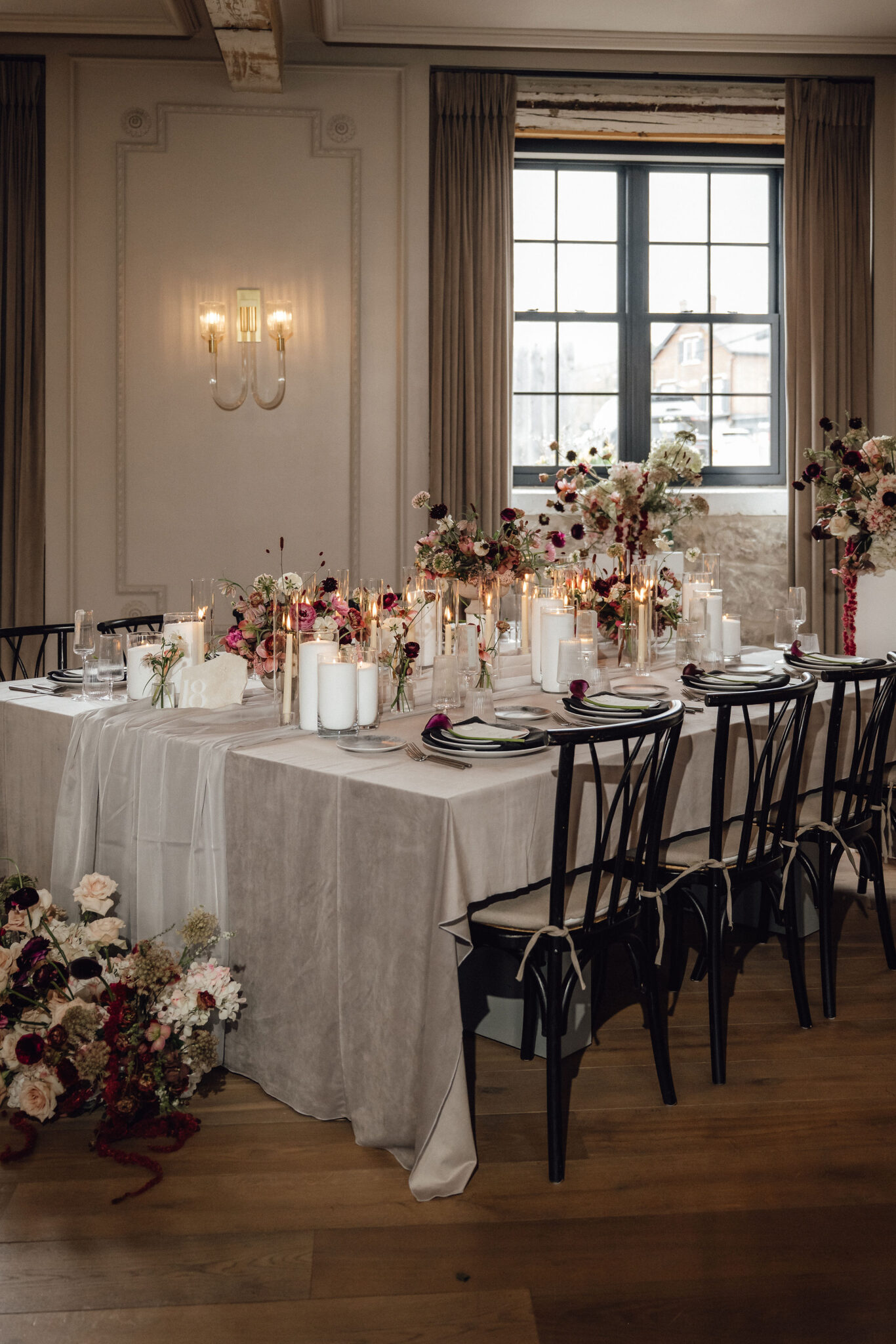 Long banquet table with white cloth, pink and white floral centerpieces, and candle votives in a softly lit dining room with a large window.