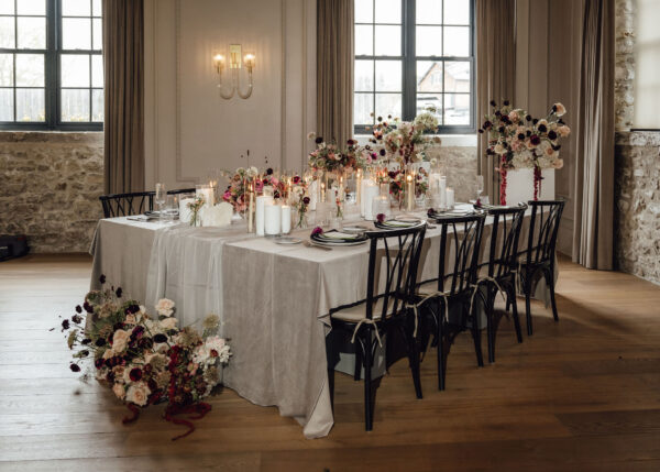 Banquet table set for a wedding with pink and white floral centerpieces and tall candles in a rustic room.