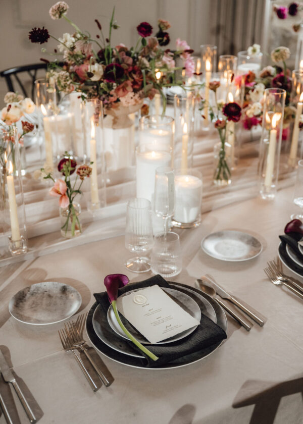 Elegant table setting with layered plates, silverware, a black napkin, and a purple flower on the napkin beside a menu card under candlelight and floral centerpiece.