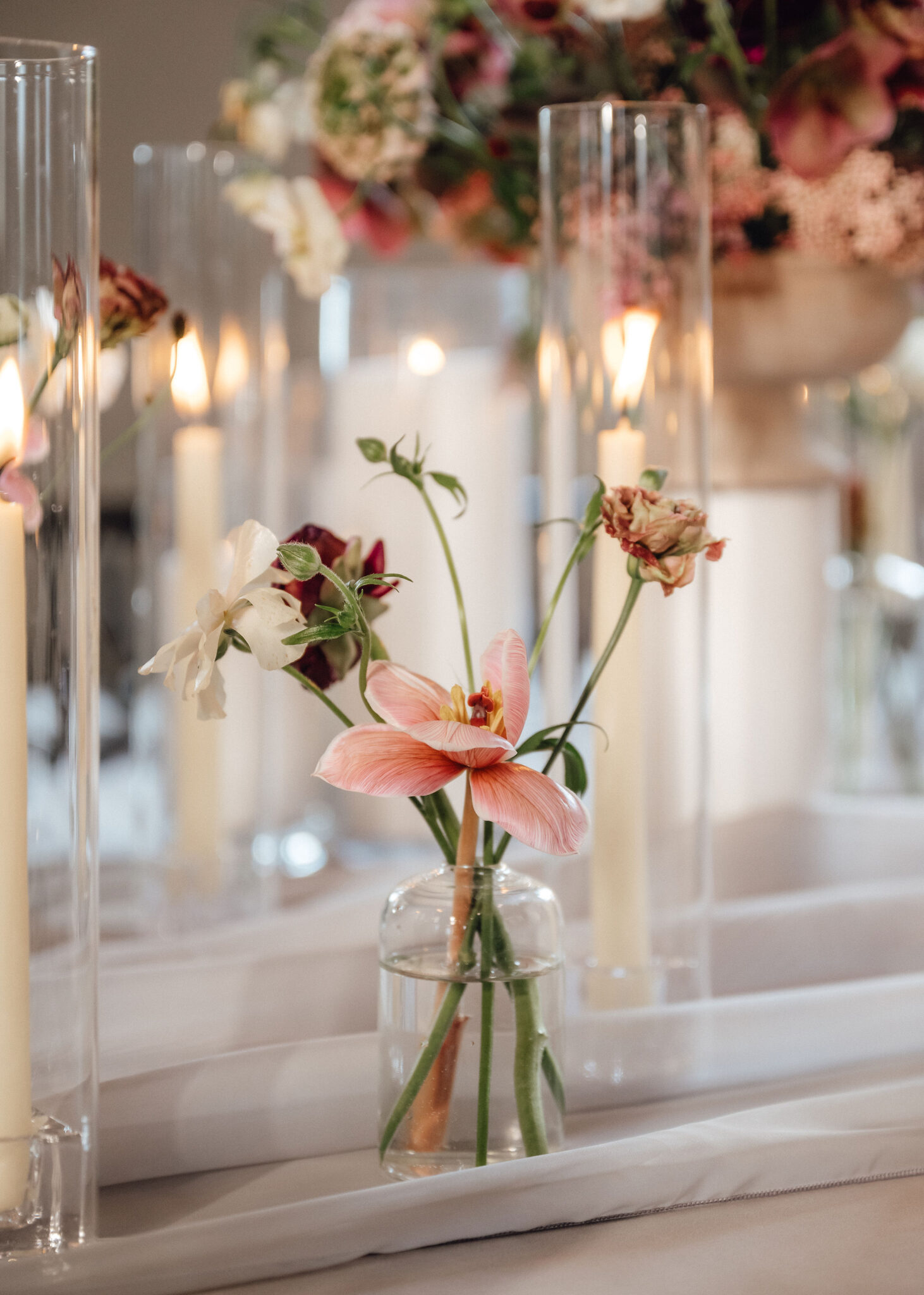 Close-up of pink flowers in a small glass vase on a white table, with tall cylindrical vases and candlelight blurred in the background.