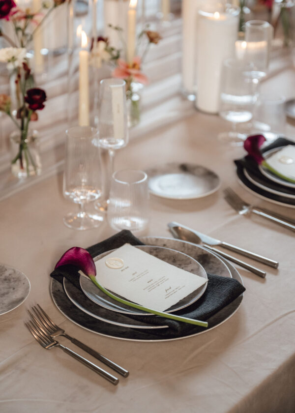 Elegant banquet table with white linens, stacked plates, crystal glasses, and a pink flower on the menu card at each setting.