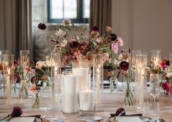 Wedding reception table with a tall glass candle centerpiece and a lush pink‑burgundy floral arrangement