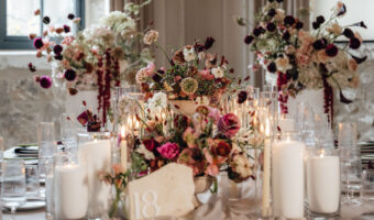 Banquet table with white linens, tall pink and white floral centerpiece, candles, and glassware in a softly lit reception setting.