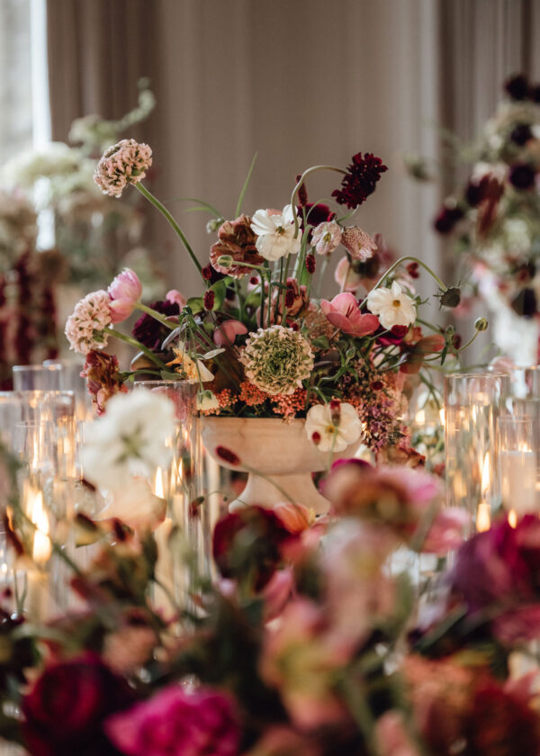 Floral centerpiece with pink, white, and burgundy blooms in a white pedestal vase, surrounded by glass candleholders and soft candlelight in a dining setup.