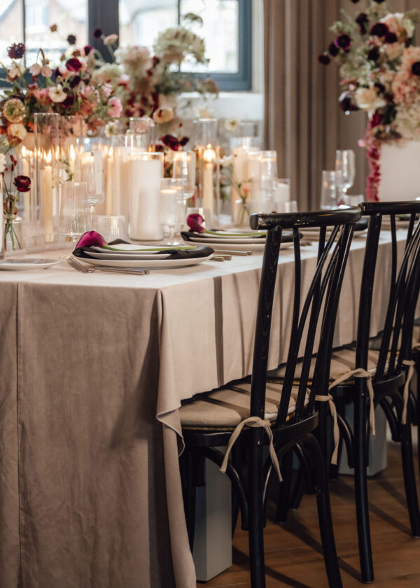 Long banquet table draped in beige cloth with candles, glassware, and tall floral centerpieces, surrounded by black wooden chairs.