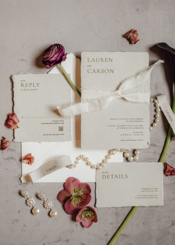 Wedding invitation suite with 'LAUREN and CARSON' and 'REPLY' cards, cream paper, white ribbon, pearls, and pink flowers.