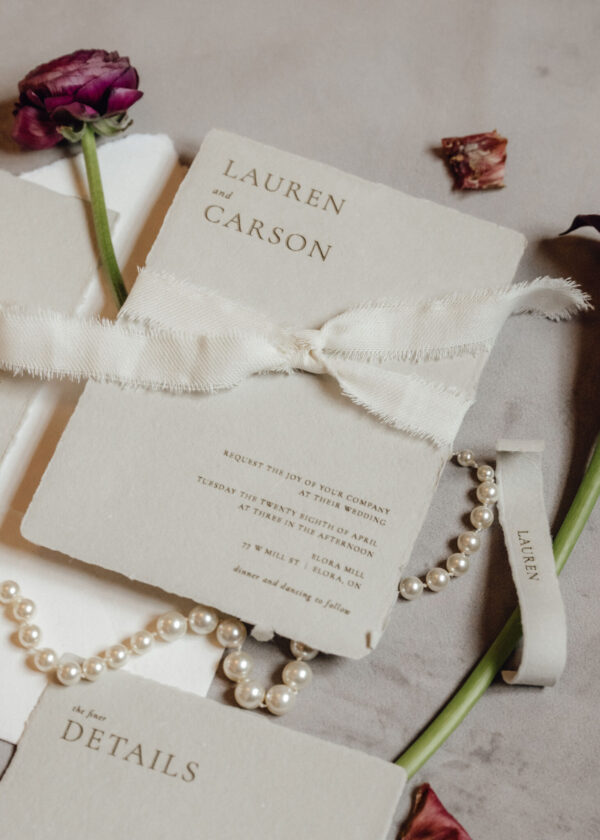Wedding invitation details card with 'DETAILS' text, tied with a white ribbon, accompanied by a pearl necklace and dried purple rose petals.