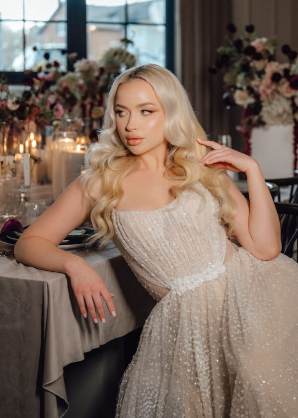 Blonde woman in a glittering champagne dress sits at a candlelit banquet table with floral decor to the background.