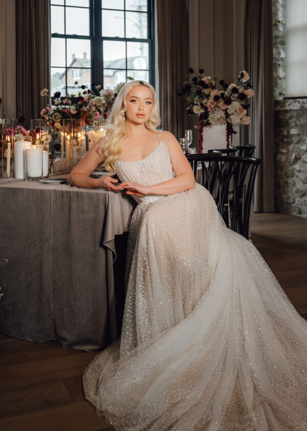 Bride in a sparkling white gown sits at a candlelit table with floral centerpieces in an elegant room by a large window.