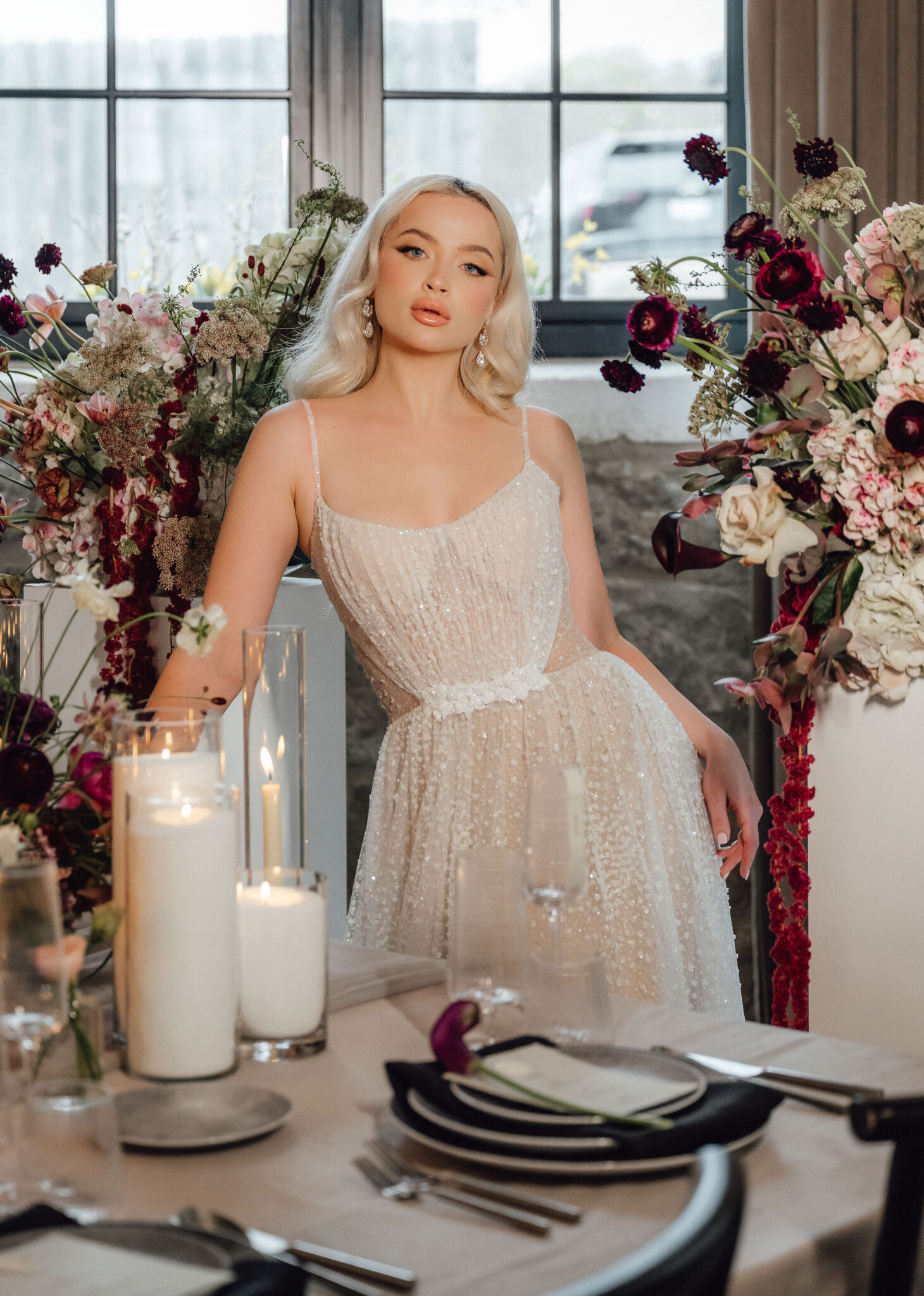 A woman in a beaded white dress sits at a table surrounded by tall floral arrangements and candles in a bright room.