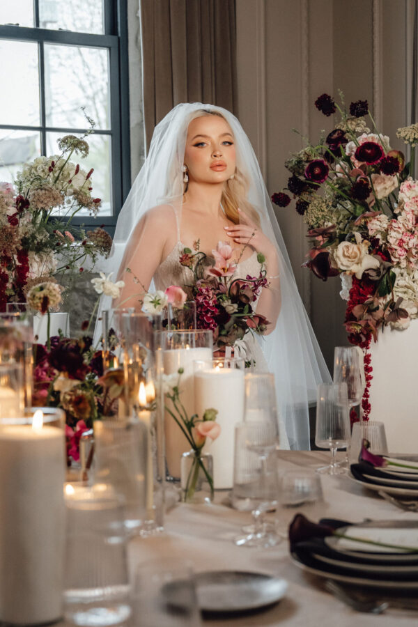 Bride in a white wedding dress and veil sits at a candlelit table surrounded by pink and burgundy flower arrangements and glassware.